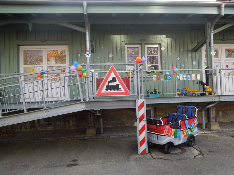 Eine Rampe führt zu einem mit Luftballons geschmückten Gebäude, vor dem ein Schild mit einem Dampfzug und zwei Wagen mit der Aufschrift "Tigerstation" stehen.