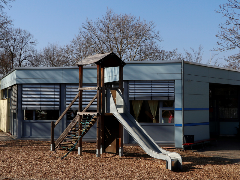 Ein moderner Spielplatz mit einer Rutsche und Klettermöglichkeiten steht vor einem flachen, grauen Gebäude.