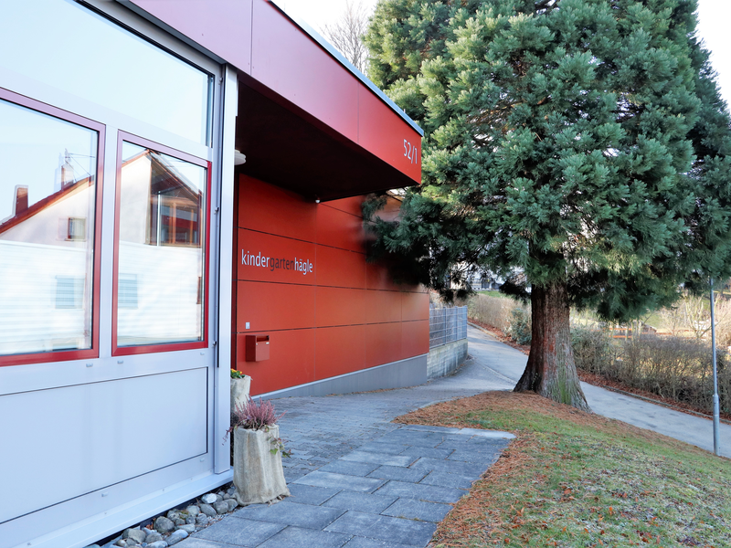 Das Bild zeigt das moderne Gebäude eines Kindergartens mit der Aufschrift "kindergarten hägle", neben einem großen Baum und einem gepflasterten Gehweg.