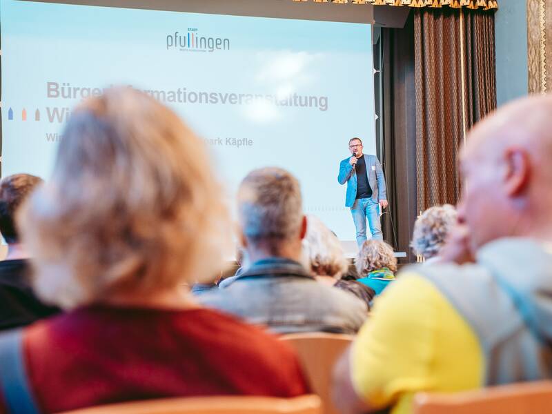 Bürgermeister Stefan Wörner steht auf einer Bühne und hält ein Mikrofon, während im Hintergrund eine Präsentationsfolie mit dem Titel "Bürgerinformationsveranstaltung Windenergie" projiziert wird.