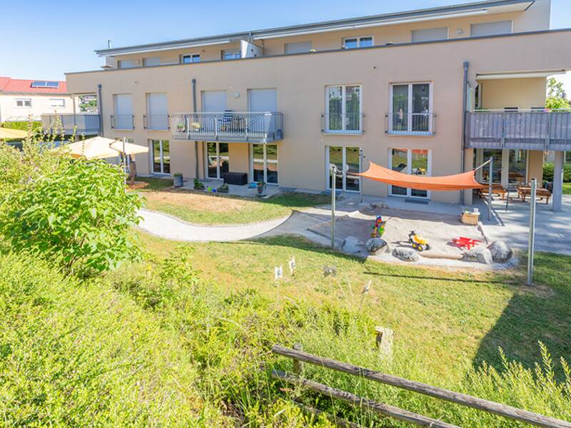 Ein helles Gebäude mit vielen Fenster, im Vordergrund ist ein Sandkasten mit orangenem Sonnenschutz in Mitten von grünen Büschen und Rasen.