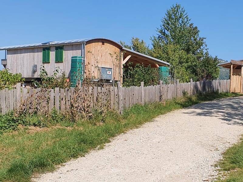 Seitlich von einem Kiesweg stehen zwei moderne hölzerne Bauwaren hinter einem Holzzaun umgeben von grünen Bäumen und Wiese.